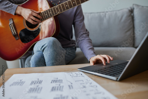 Closeup of young woman using laptop while playing guitar at home, online music lesson, copy space