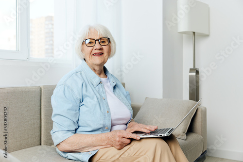 a nice, sweet elderly lady is sitting on a beige sofa resting and smiling pleasantly looking at the camera holding a laptop on her lap, keeping herself up to date with new events