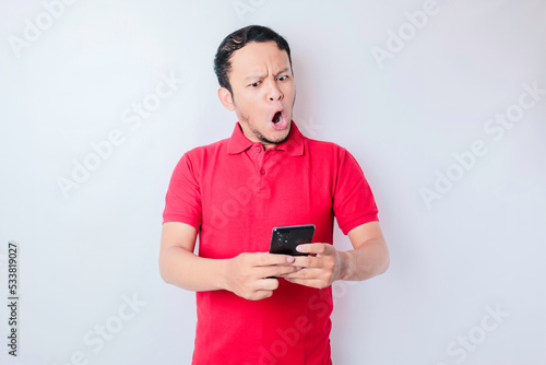 Surprised Asian man wearing red t-shirt pointing at his smartphone, isolated by white background © Reezky