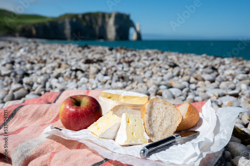 Lunch with french round camembert cow cheese from Calvados region, fresh baked baguette bread and apple on pebbles stones beach with view on alebaster cliffs Porte d'Aval in Etretat, Normandy, France