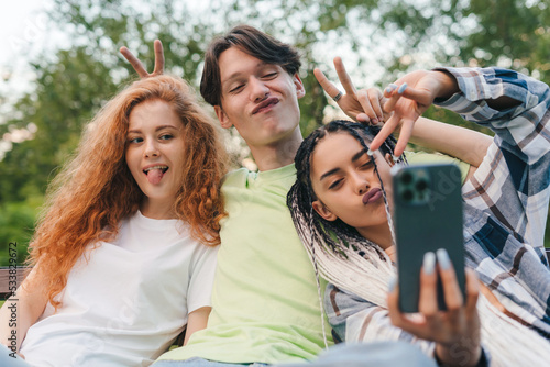 A group of friends taking a selfie with a smart phone sitting on a bench in the park looking the camera making funny faces. School education. Mobile phone