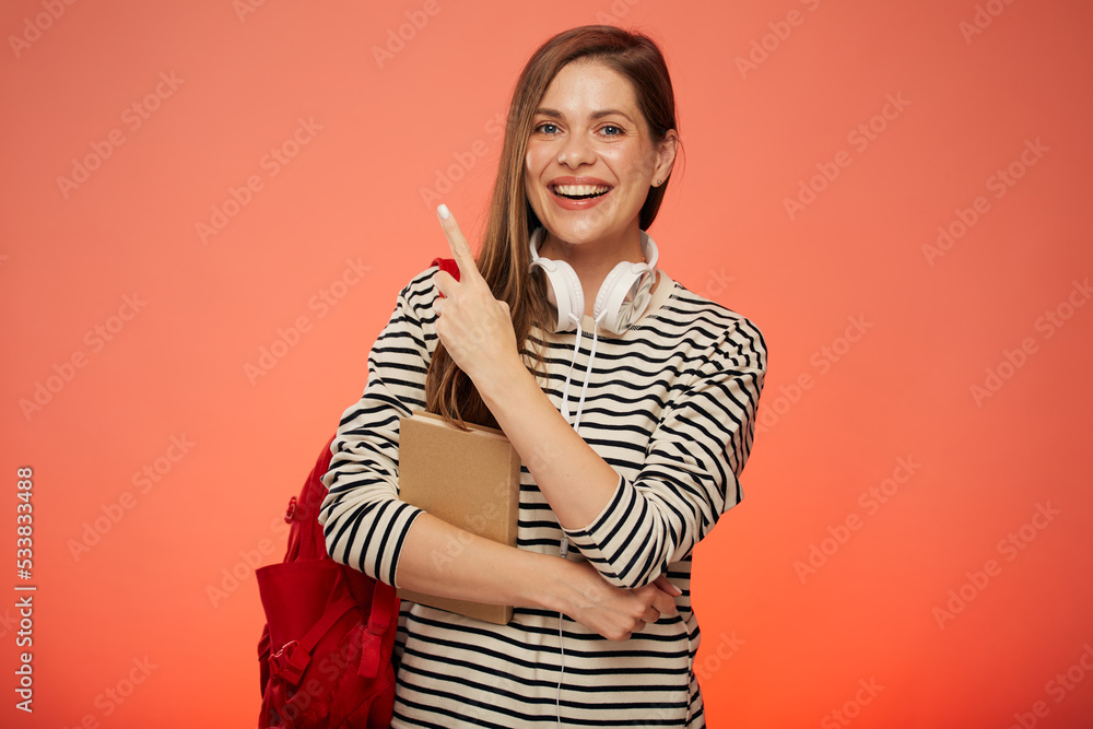 Obraz premium Smiling student with book and red backpack pointing finger away. isolated female portrait on pink background.