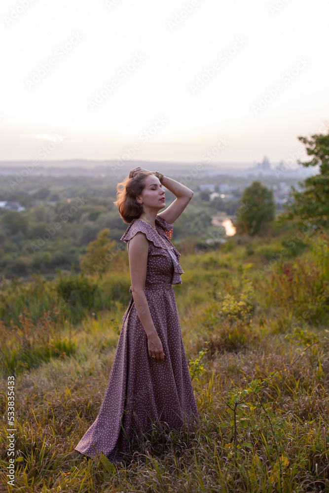 Beautiful young smiling girl in a long brown dress stands along the lawn. Happy woman walks at sunset on a hill overlooking the river. Concept of having rest in park during summer holidays or weekends Beautiful young smiling girl in a long brown dress stands along the lawn. Happy woman walks at sunset on a hill overlooking the river. Concept of having rest in park during summer holidays or weekends