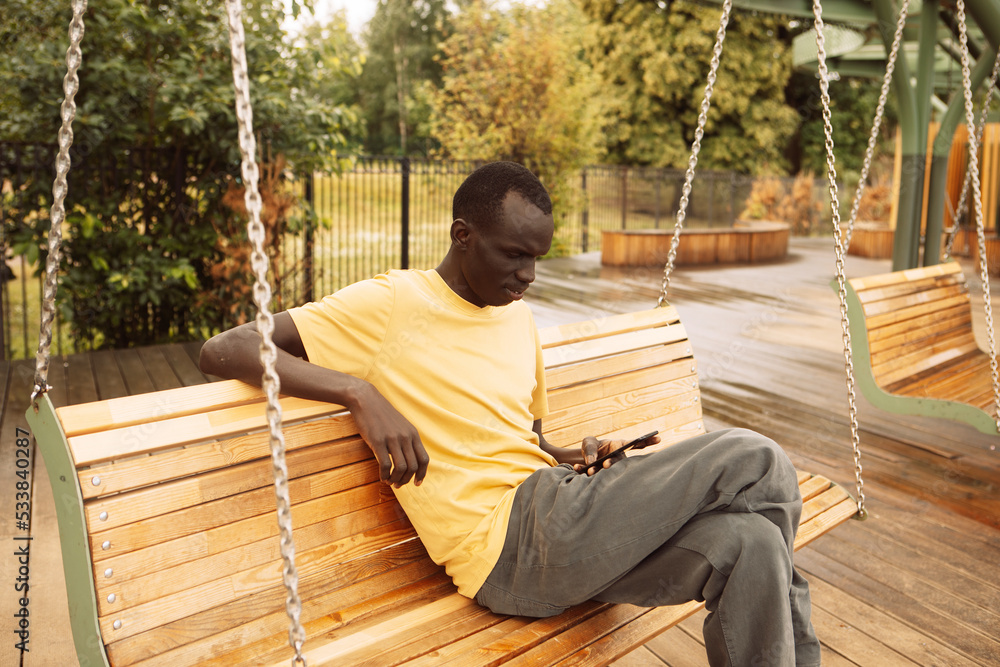 Dark-skinned afro american young man sitting on a swing in the park on a summer day, holding a smartphone and texting or browsing the news on social networks. Dark-skinned afro american young man sitting on a swing in the park on a summer day, holding a smartphone and texting or browsing the news on social networks.