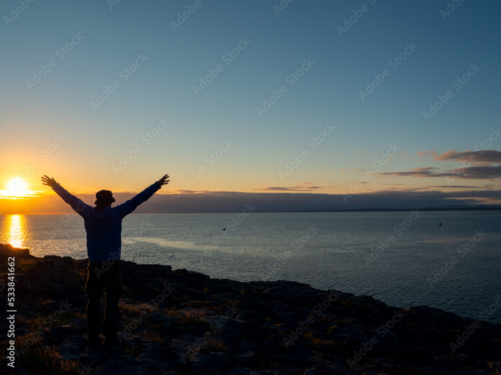 Silhouette of a slim man with hands up in the air. Stunning sunset scene in the background with dramatic sky and Atlantic ocean. Galway bay, Ireland. Emotion expression through body language.
