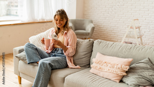 Happy woman drinking coffee in early morning, sitting on sofa in pink  shirt at home. Using smartphone.