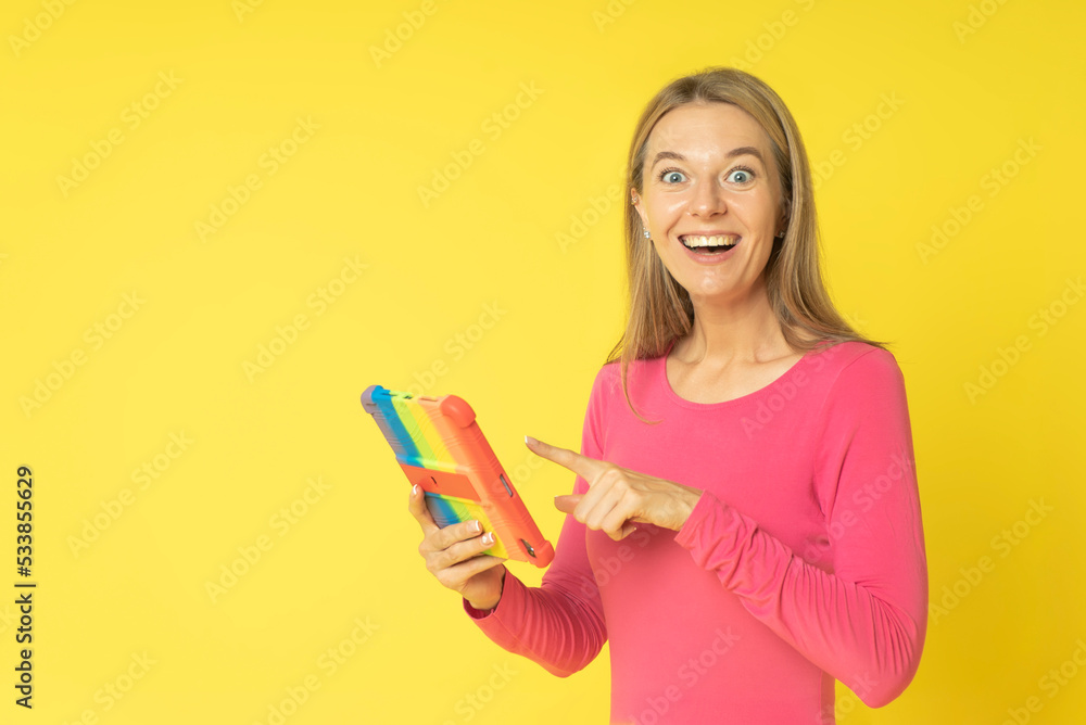 Young smiling happy fun woman 30s wearing pink dress holds tablet computer in colored cover pointing index finger isolated on yellow color background studio portrait. Young smiling happy fun woman 30s wearing pink dress holds tablet computer in colored cover pointing index finger isolated on yellow color background studio portrait.