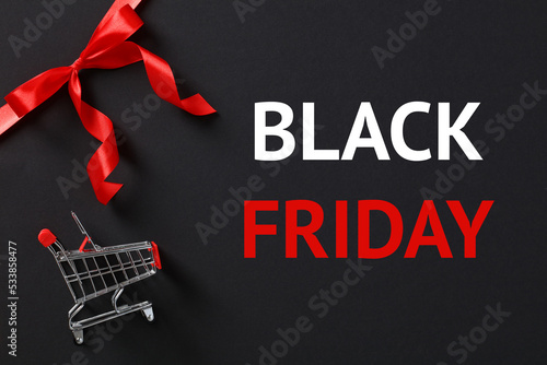 Black friday sale concept with red ribbon bow in corner and shopping cart on dark background.