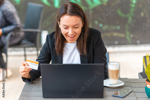 Young smiling business woman holding in hand credit bank card and paying online while sitting at cafe table with laptop, sitisfied female office employee shopping buying goods in internet. photo