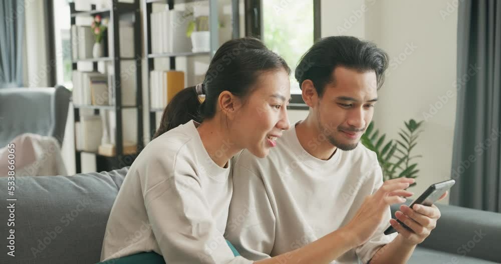 Happy young Asian couple sit on couch have fun use smartphone devices online shopping together at home. Smiling young husband and wife laugh relax at home browsing application on mobile phone gadgets. Happy young Asian couple sit on couch have fun use smartphone devices online shopping together at home. Smiling young husband and wife laugh relax at home browsing application on mobile phone gadgets.
