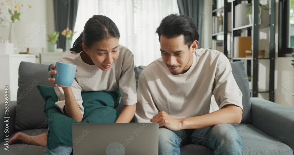 Happy young Asian couple sit on couch fun use laptop computer online shopping at home on holiday. Smiling young husband and wife laugh relax at home browsing application on gadgets, Lifestyle concept. Happy young Asian couple sit on couch fun use laptop computer online shopping at home on holiday. Smiling young husband and wife laugh relax at home browsing application on gadgets, Lifestyle concept.