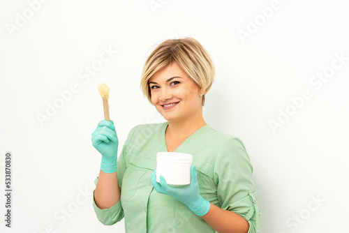 The master of sugar hair removal holds liquid yellow sugar paste, wax for depilation with the jar on a wooden stick on a white background photo