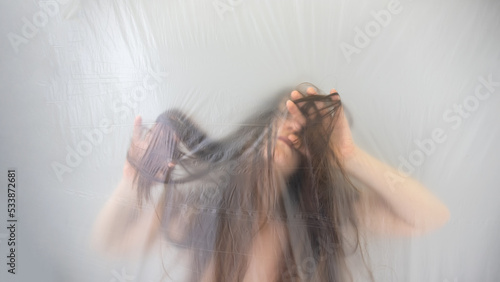 blurred, fuzzy image of sensual romantic, young woman moving in trance, playing with her long hair, behind transparent plastic foil film