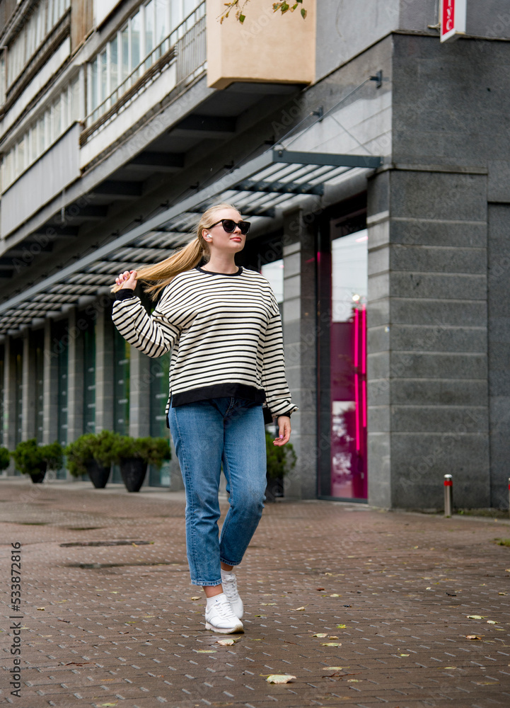 smiling woman with long blond hair in sunglasses and blue jeans walking on the street