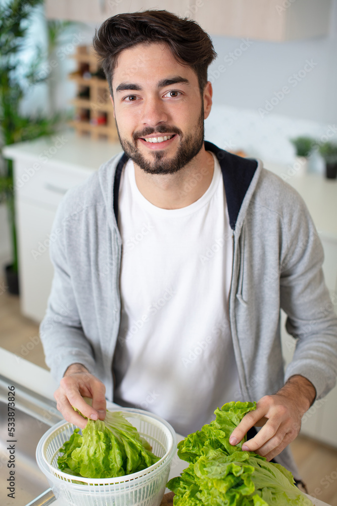 Fototapeta premium glad young bearded man standing at kitchen and preparing salad