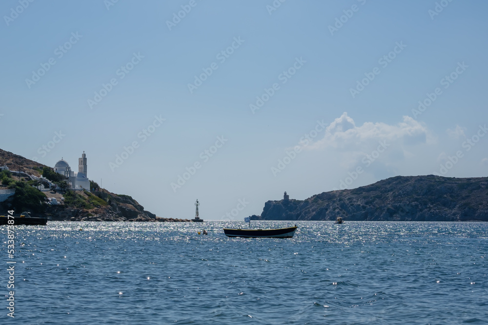 View of a little fishing boat with a Greek flag, a beautiful church and the lighthouse in the background at the port of Mylopotas in Ios Greece