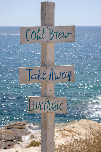 View of wooden signs for Take away, Cold brew, Live music and the sea in the background on the beach of Mylopotas in Ios Greece