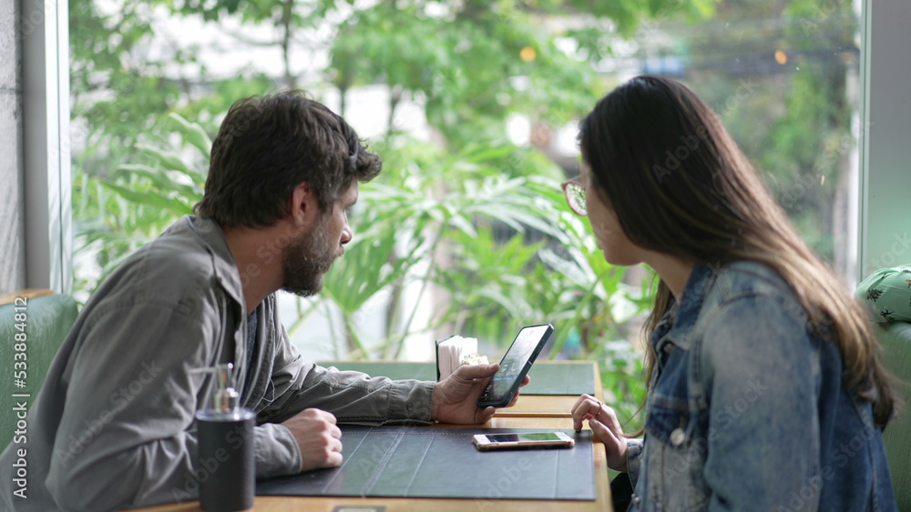 Candid couple sitting at coffee shop looking at smartphone device screen. Man and woman staring at phone seated at cafe restaurant Candid couple sitting at coffee shop looking at smartphone device screen. Man and woman staring at phone seated at cafe restaurant
