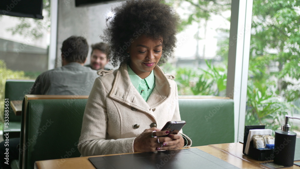 One black woman checking smartphone device sitting at coffee shop table. An African American girl browsing internet on phone One black woman checking smartphone device sitting at coffee shop table. An African American girl browsing internet on phone