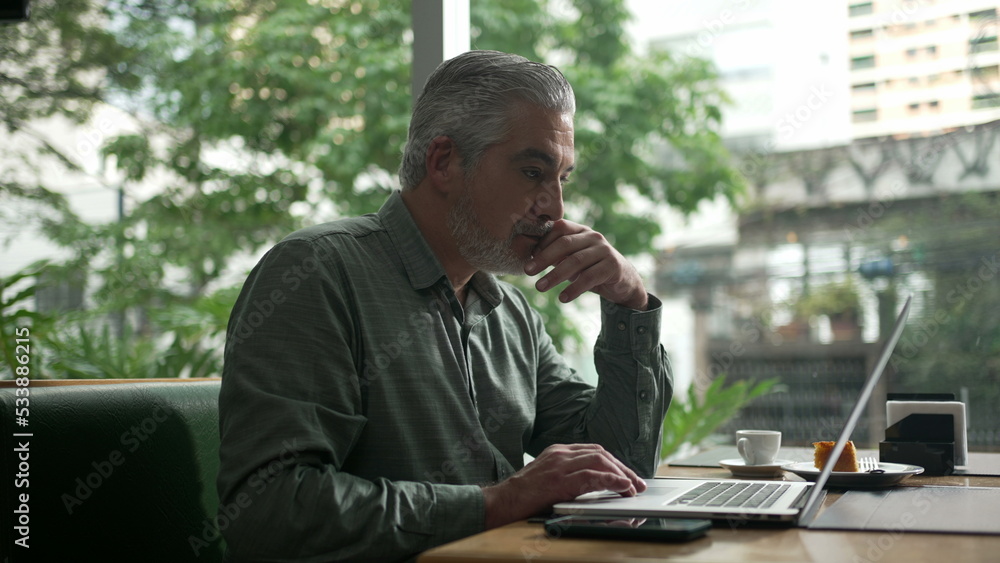 Middle aged man working at coffee shop in front of laptop. Older person receiving message notification on phone. Executive person typing on computer Middle aged man working at coffee shop in front of laptop. Older person receiving message notification on phone. Executive person typing on computer