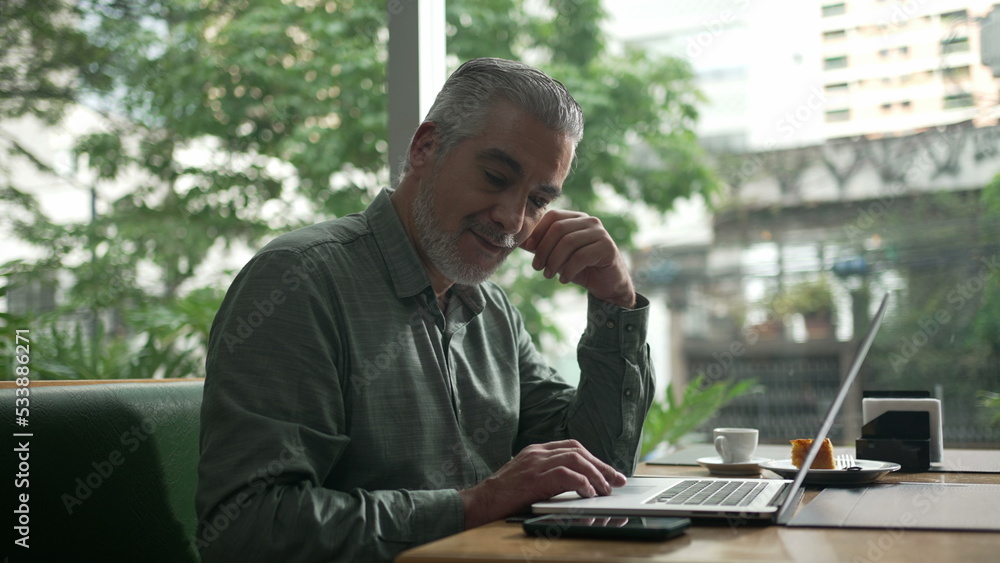 Middle aged man working at coffee shop in front of laptop. Older person receiving message notification on phone. Executive person typing on computer Middle aged man working at coffee shop in front of laptop. Older person receiving message notification on phone. Executive person typing on computer