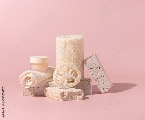 Face towel, loofah sponges and cream jar on stones on light beige close up. Mockup