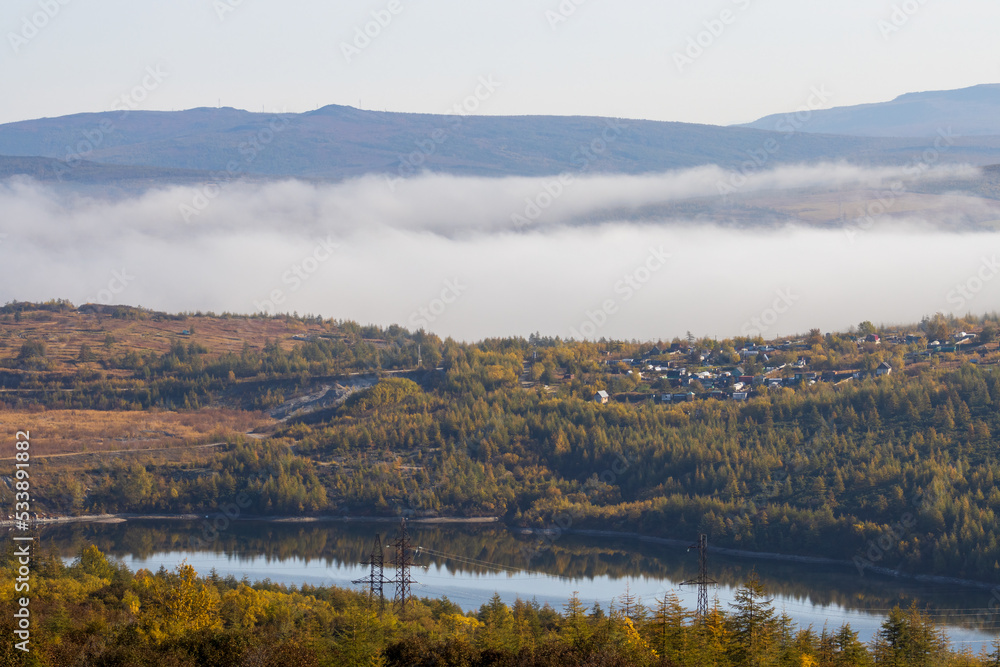 Foggy rural landscape. View of the village among the hills. In the background, fog in the mountains. Reservoir in the foreground. Magadan region, Siberia, Russian Far East. Autumn season, September.