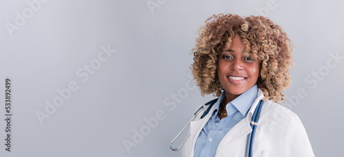 Smiling mixed race doctor in a and white coat, holding a stethoscope on a gray background looking at the camera, baner. Concept: doctor, medicine, practice, treatment of diseases