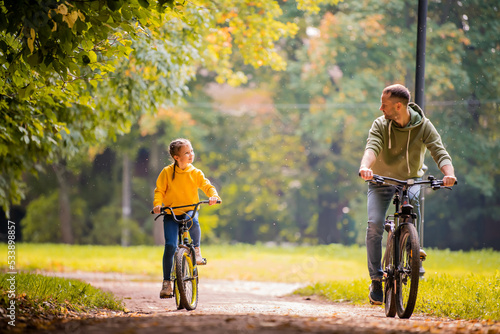 Happy father and daughter ride bicycles in autumn park on sunny day.