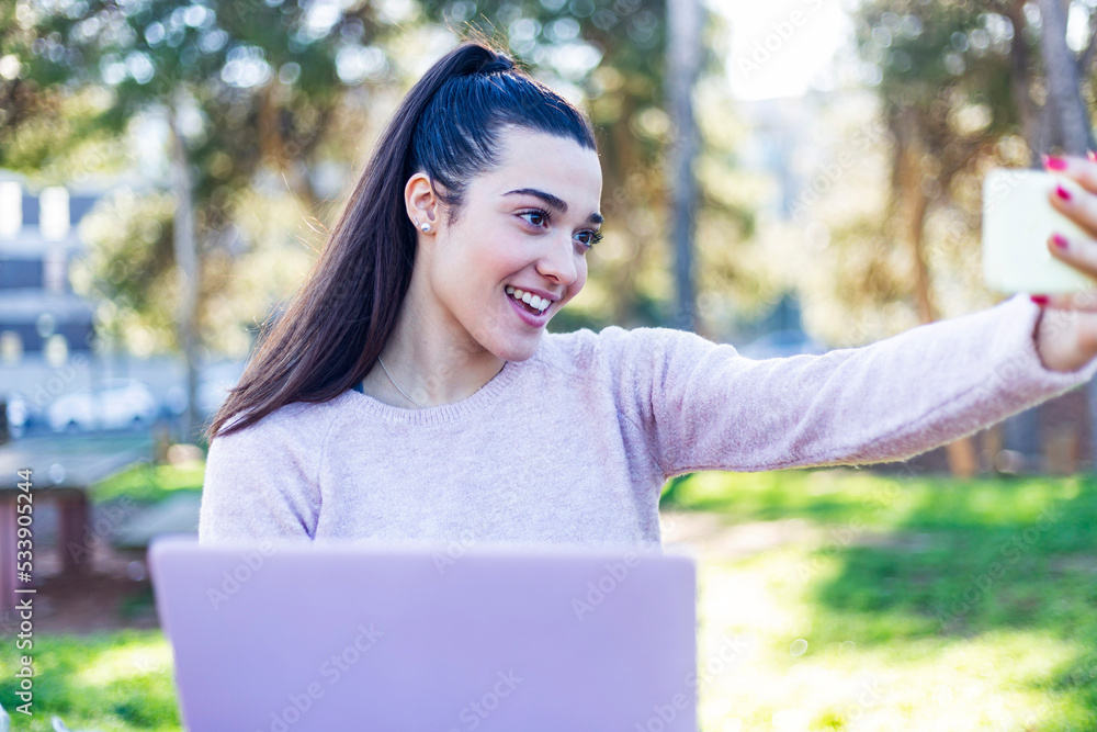 charming girl with a laptop makes a selfie while sitting on a bench in the park charming girl with a laptop makes a selfie while sitting on a bench in the park