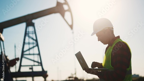 oil business. a worker works next to an oil pump holding a laptop. industry business oil and gas concept. lifestyle engineer studying the level of oil production on a laptop silhouette at sunset