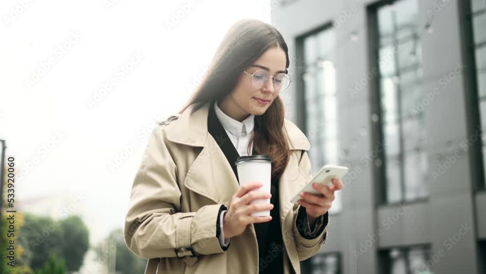 Charming young office worker in eyeglasses have a coffee break using social media application on smartphone, texting messages, receive news near business centre. Charming young office worker in eyeglasses have a coffee break using social media application on smartphone, texting messages, receive news near business centre.