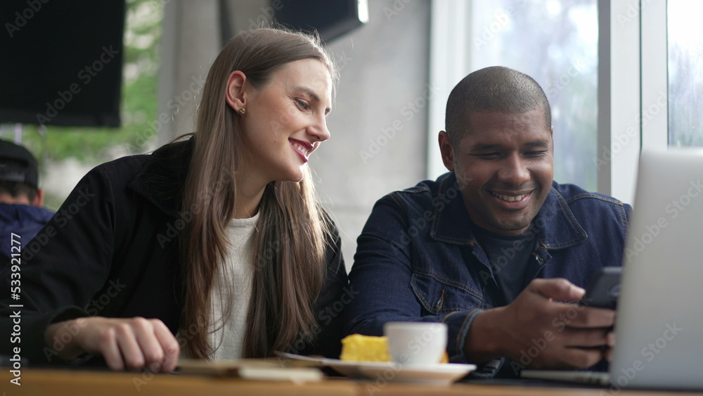 Young diverse couple seated at coffee shop looking at phone screen together. Happy people using smartphone sharing screen. Authentic real life laugh and smile Young diverse couple seated at coffee shop looking at phone screen together. Happy people using smartphone sharing screen. Authentic real life laugh and smile