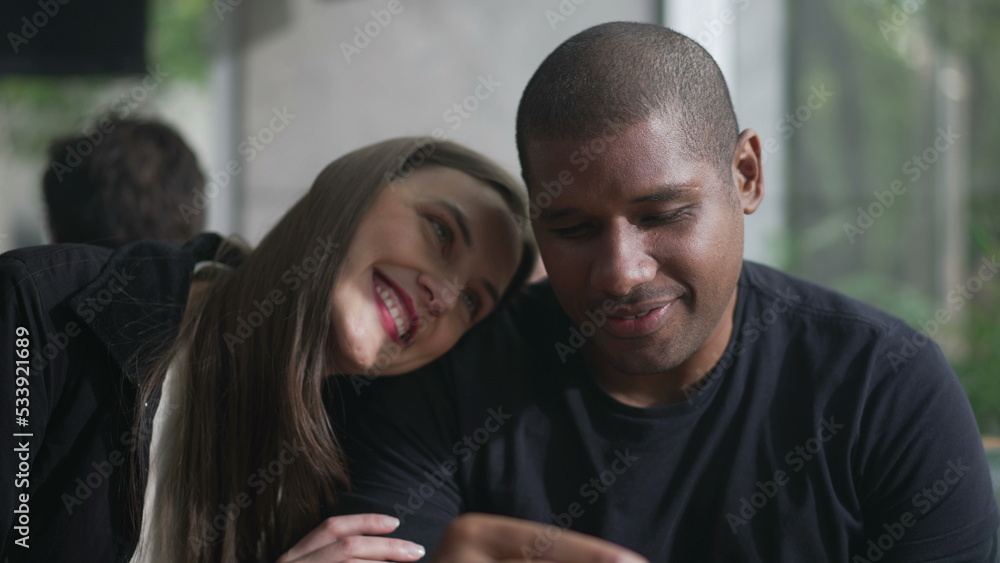 White girl leaning on a black man shoulder. A diverse couple smiling seated at coffee shop. Boyfriend and girlfriend on a romantic date