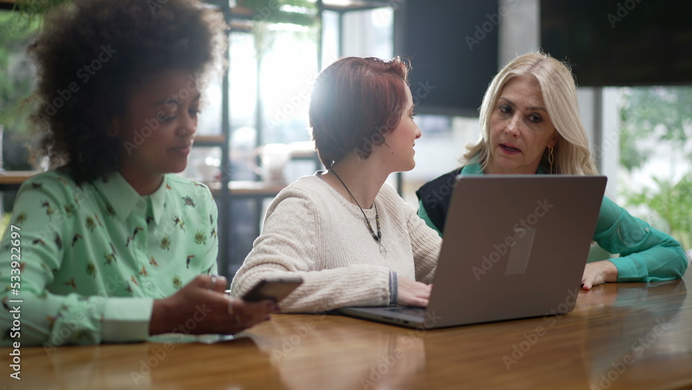 Three diverse women in front of laptop seated at coffee shop. Young employees with business woman executive coaching employees Three diverse women in front of laptop seated at coffee shop. Young employees with business woman executive coaching employees