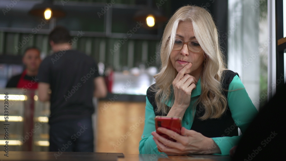 Senior female executive woman using smartphone at coffee shop. Undecided older person browsing internet online seated at cafe place restaurant Senior female executive woman using smartphone at coffee shop. Undecided older person browsing internet online seated at cafe place restaurant