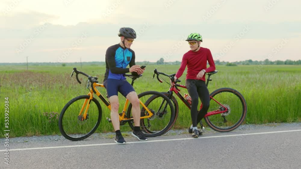 Male athletes sitting on bikes and resting during intensive training for international competition. Cycling man in helmet and glasses checking his coordinate on smartphone. Male athletes sitting on bikes and resting during intensive training for international competition. Cycling man in helmet and glasses checking his coordinate on smartphone.