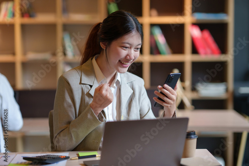 Beautiful Asian businesswoman using smartphone is delighted and excited by the success she has achieved through her office cell phone.