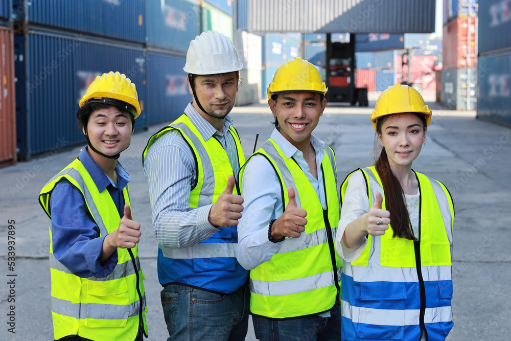 Group of multiethnic technician engineer or worker in protective uniform with hardhat standing and showing thumb up celebrate successful together or completed deal commitment at container cargo site Group of multiethnic technician engineer or worker in protective uniform with hardhat standing and showing thumb up celebrate successful together or completed deal commitment at container cargo site