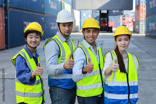 Group of multiethnic technician engineer or worker in protective uniform with hardhat standing and showing thumb up celebrate successful together or completed deal commitment at container cargo site