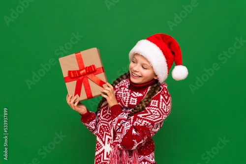 A beautiful girl in a Christmas sweater and a Santa Claus hat is holding a New Year's gift in her hands. A child with pigtails and a warm winter sweater with a gift in his hands.