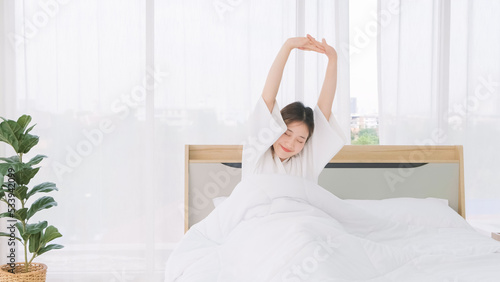 Asian woman wearing bathrobe stretching her arms above her head while lying on bed. Morning relaxation routine after waking up. Woman tourist stretching her arms after waking up in bed of hotel room.