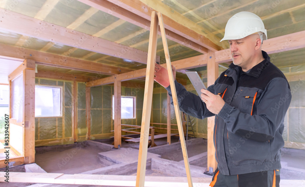 Builder inside building. Man builder with boards and tablet. Decorating house with wood. Tablet in hands of builder. Concept using building apps. Construction worker stands in unfinished house