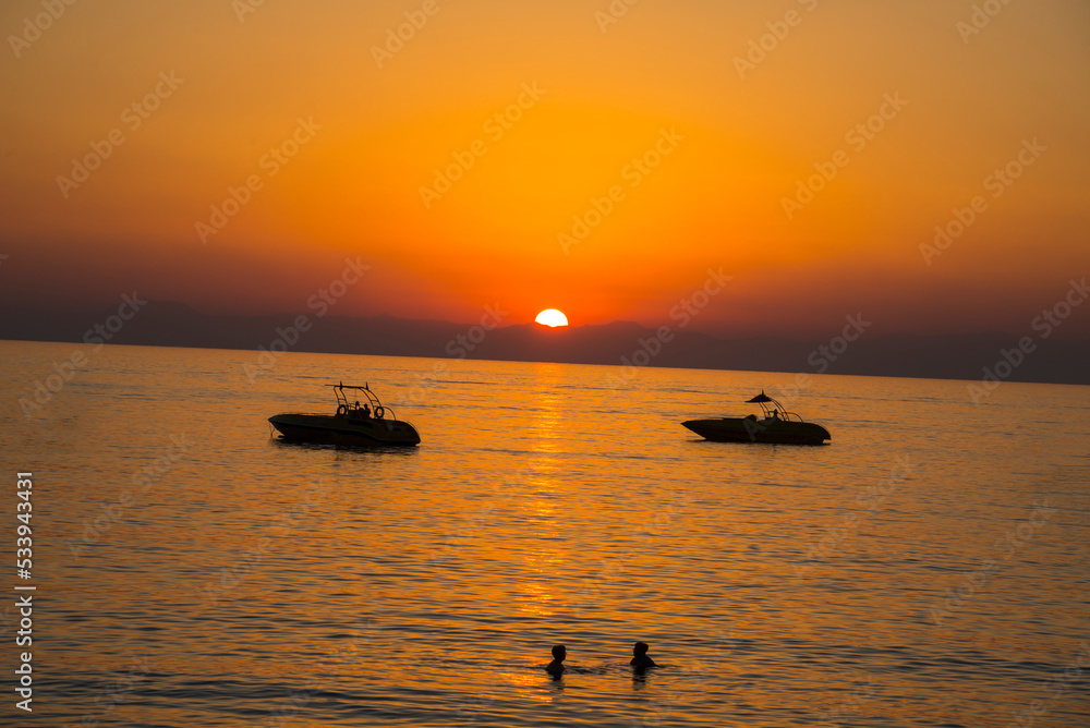 Sunset at the beach with boats and silhouette of swimming people.