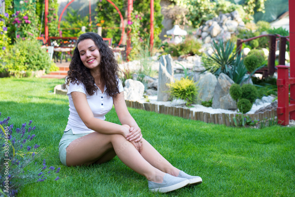 Young smiling woman sitting on the green grass of her garden on a beautiful summer day Young smiling woman sitting on the green grass of her garden on a beautiful summer day