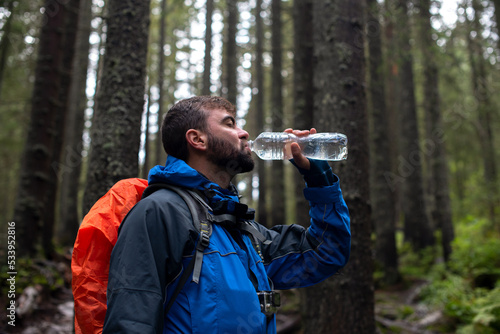 Young attrective male hiker with beard drinking water from plastic bottle standing in a mountain green forest. Acitivity and sport concept. Thirsty man. Tourism background. Copy space