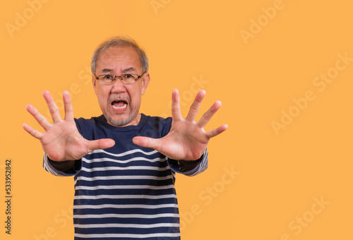 Portrait of Asian senior man wearing glasses standing face expression and two hands rising posing stop symbol isolated on yellow background.