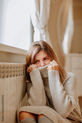 Young woman in long winter beige sweater is posing at home near the radiator