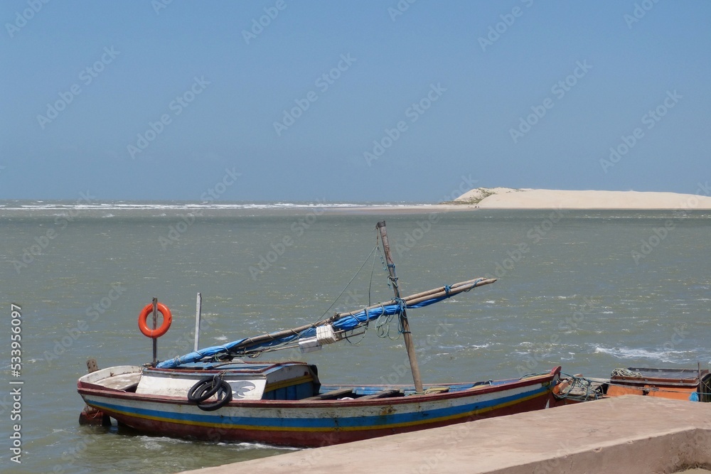 Small wooden sailboat moored at a pier in a bay and in the background a sand dune in São Luís, Maranhão, Brazil.