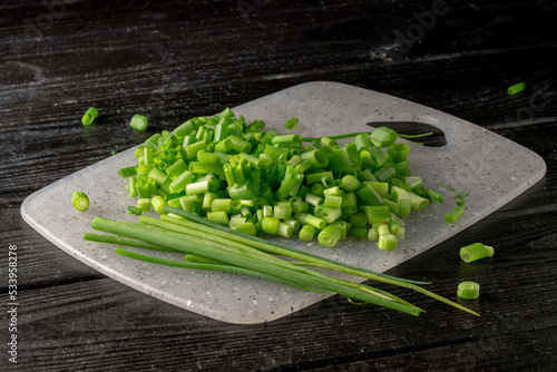 Cutting pieces and of green onion stalks on gray plastic board. Slices and stems of fresh green scallion on dark gray wooden table. Chopped shallots or chive, seasonal greenery, seasoning. Close up.